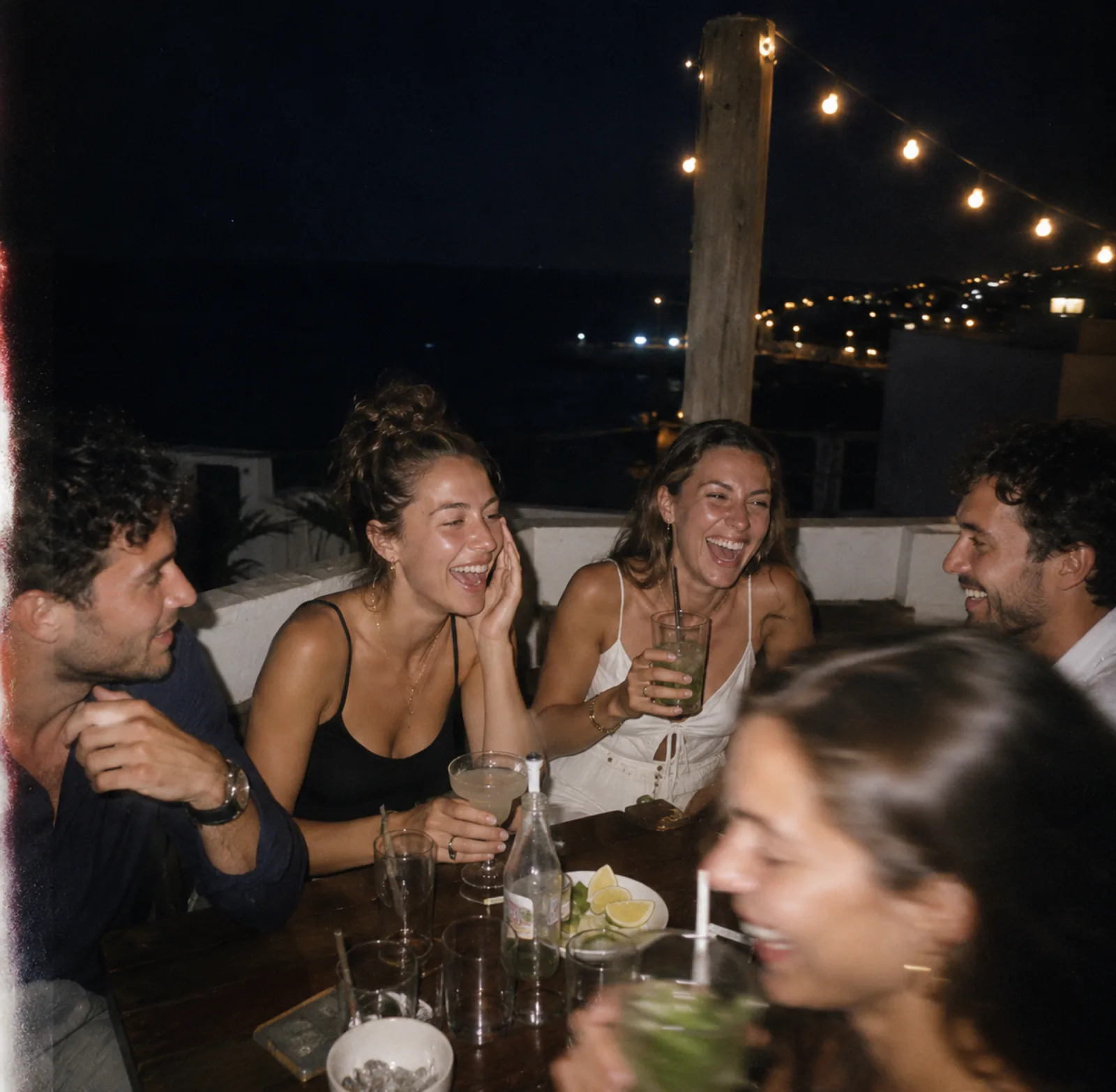 Dîner rooftop nocturne au bord de mer, cinq amis riant sous une guirlande lumineuse