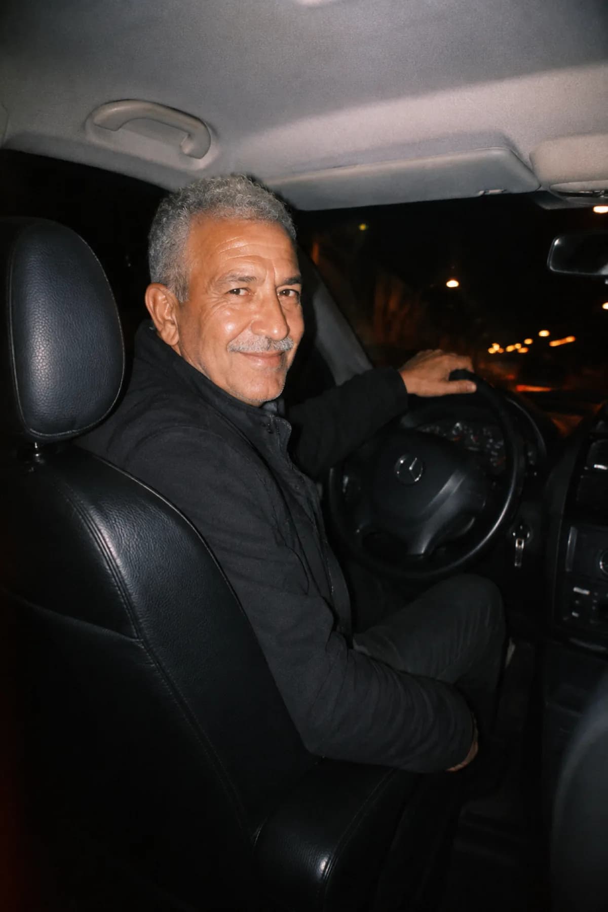 Hassan, le doyen des chauffeurs SDM, cheveux gris et sourire bienveillant au volant de sa Mercedes