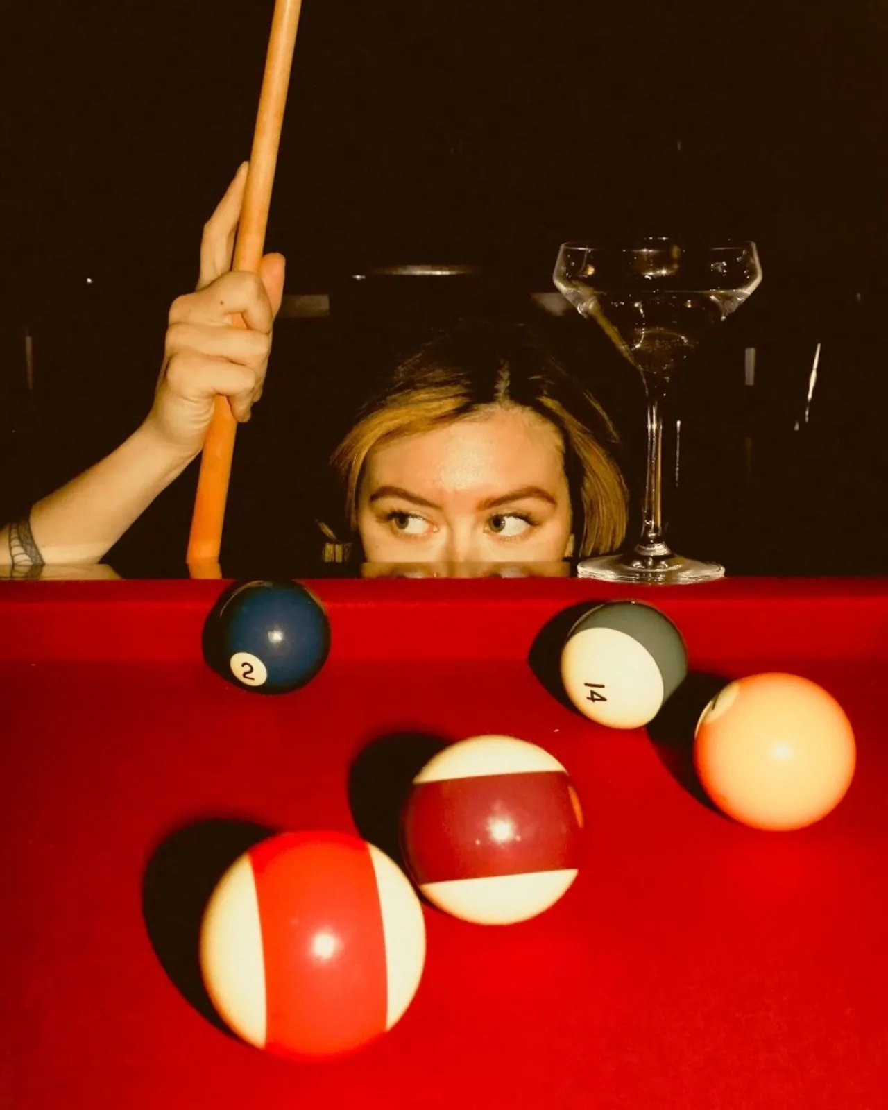 Jeune femme derrière un billard au tapis rouge, queue à la main et coupe de cocktail à côté