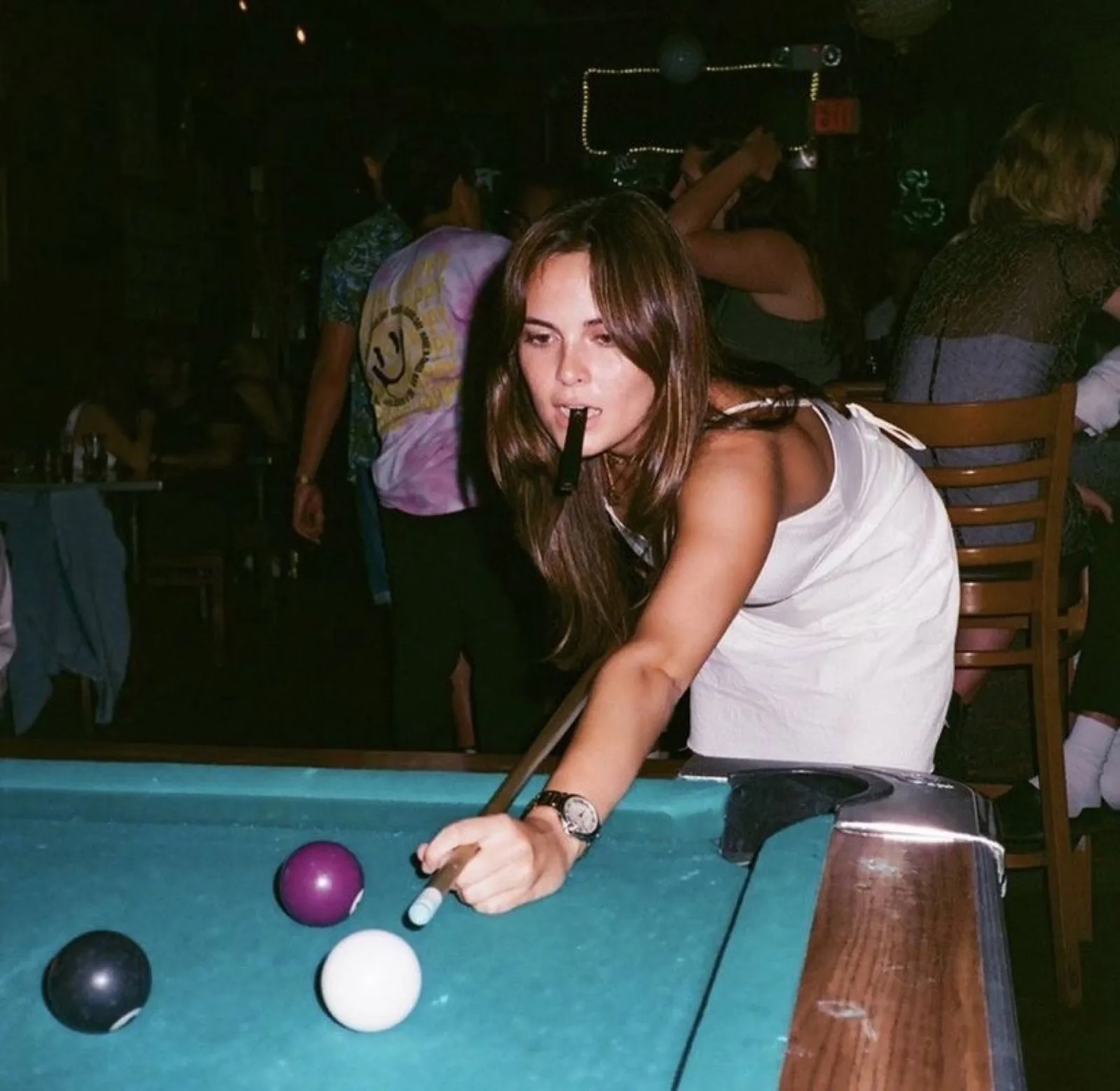 Une joueuse penchée sur un billard au tapis turquoise, queue en main, bar bondé derrière