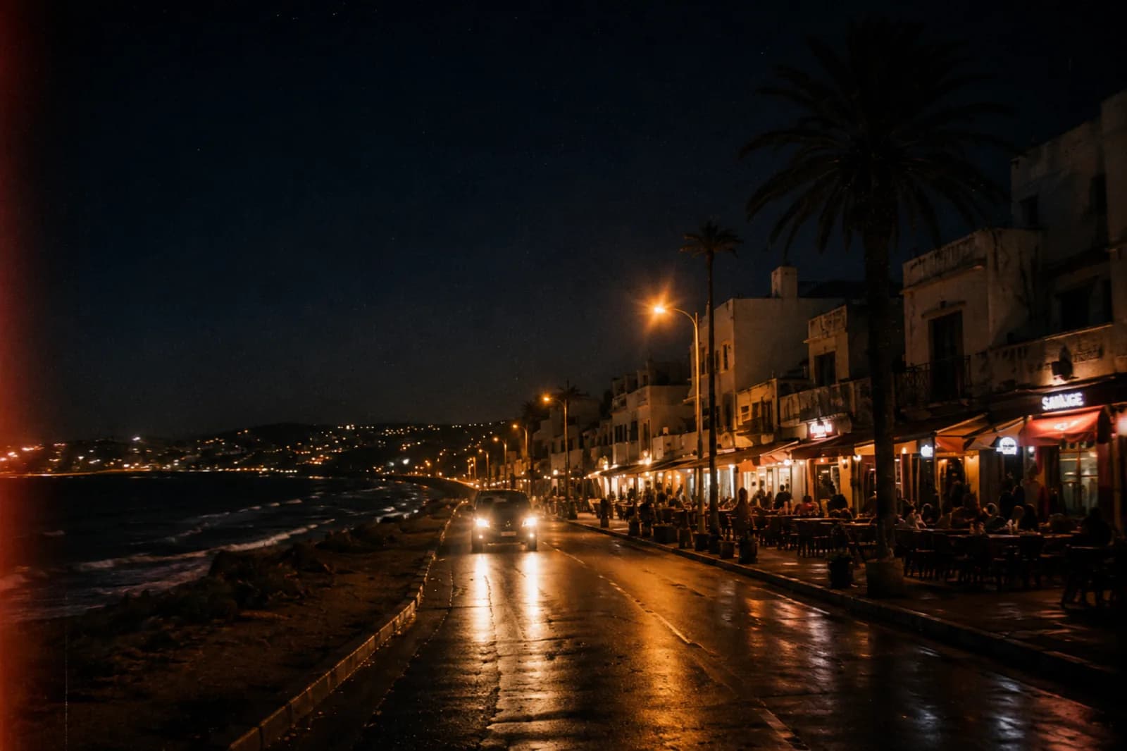 Route côtière marocaine la nuit, phares d'une voiture, terrasses de cafés animées et océan