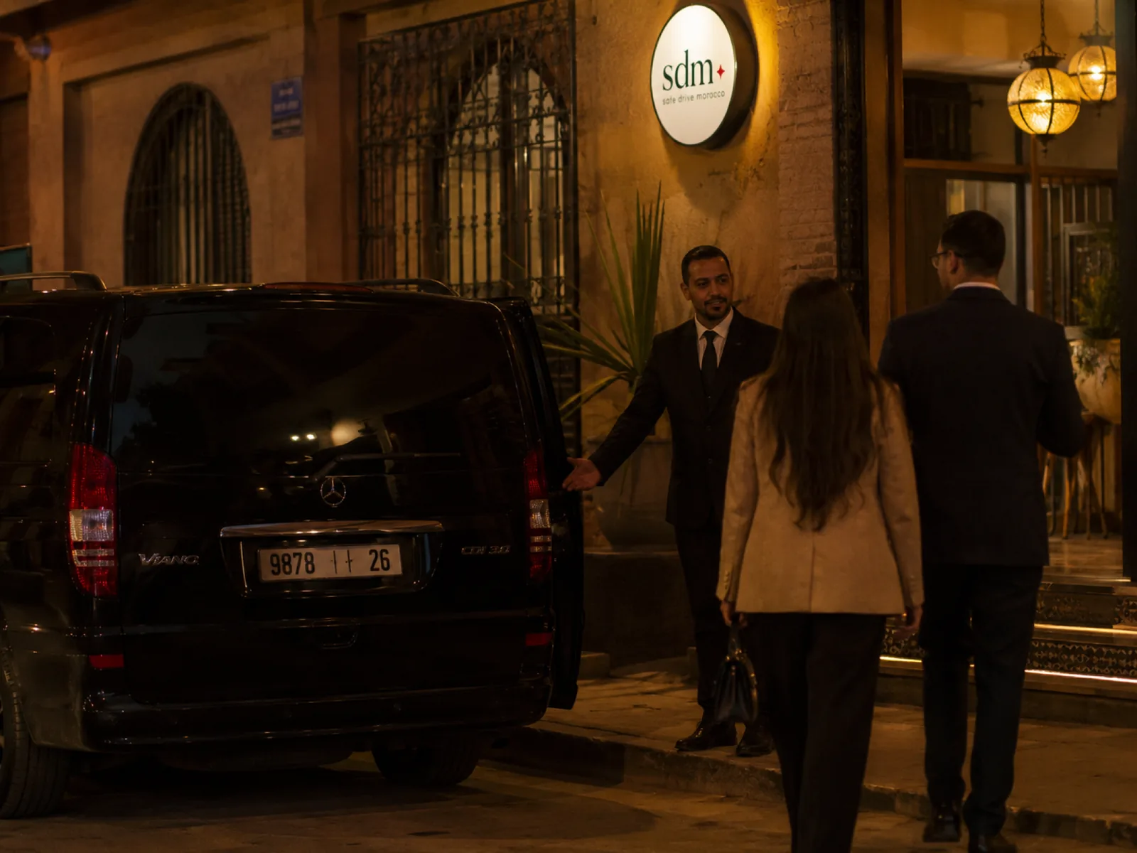 Voiturier SDM ouvrant la portière du Viano à un couple élégant devant un restaurant, enseigne SDM rétroéclairée
